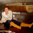 Pixelated image of a person in white shirt and dark pants seated on wooden bench with orange object, background blurred brown tones.