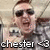 Pixelated image of a person with short brown hair wearing dark sunglasses and a gray jacket, holding a white object near their face.