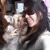 A person with long dark hair and bangs wearing a light gray top, holding a small dog in their arms while smiling.