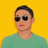 Pixelated portrait of a person with short dark hair wearing dark sunglasses and a teal shirt against a solid yellow background.