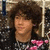 A person with brown curly hair wears a black and white patterned top with pink heart shapes, positioned against a blurred background.