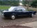 Static pixelated image of a dark blue sedan with silver accents on a road, background shows greenery and blurred structures.