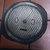 Pixelated circular icon with a gray face showing a neutral expression, dark eyes, and a straight mouth, set against a blurred background with brown and pink tones.