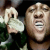 Pixelated static image of a person with light skin tone wearing dark clothing, holding an object resembling a microphone or small device near their mouth against a dark background.