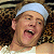 Pixelated static image of a person with light skin wearing a white head covering and open mouth expression against dark background.