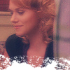 Static close-up of a person with curly red hair wearing a dark top, visible from shoulders up against an indoor background with blurred elements and pixelation at the bottom.