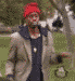 A person in a red beanie, light gray jacket, and dark blue shirt with a white emblem stands in a grassy park with trees and a white horse in the background.