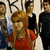 A static image of a group of people with one person in the foreground wearing a red top and long hair, others behind in dark clothing.