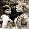 Two women in vintage-style clothing face each other against a blurred background with visible foliage.