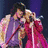 A person in a red outfit sings into a microphone while another person in purple stands behind, with a third figure visible on the right side of the frame.