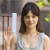 A person with shoulder-length brown hair wearing a light-colored sleeveless top, holding a white cylindrical object in their right hand against a blurred background.