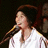 Pixelated image of a person singing into a microphone, wearing a white shirt with dark hair and brown headband against dark background.
