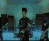 Animated sequence of a person playing electric guitar on a stage with blue lighting and band equipment in background.