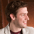 Pixelated profile view of a person with dark curly hair wearing a white shirt over a dark red garment against a brown background.