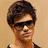Pixelated image of a person with short brown hair and dark sunglasses against a beige background.