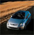 A blue sedan with a white front bumper is positioned on a dark surface beneath a brown textured background.