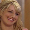 A close-up of a smiling person with blonde hair, wearing a pink garment and hoop earrings, against a blurred indoor background.