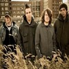 Four people stand in a row against a brick wall background with a patterned foreground, wearing dark hooded jackets and casual clothing.