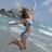 Pixelated image of a person in a blue swimsuit jumping in water with arms raised, background shows sky and water.