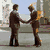 Two men in brown suits shake hands on a city street with buildings and a statue in the background.