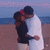 Two people embrace on a beach at sunset, with the ocean and sky in the background.