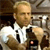 A pixelated image shows a person with light hair wearing a white shirt with dark accents, holding a dark object near their chest against a blurred background.
