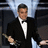 A man in a black tuxedo with white bow tie holds an award, standing against dark chairs and light blue backdrop.