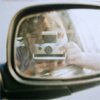 A side-view mirror reflects a person holding a silver digital camera with visible buttons and screen, set against blurred background elements.