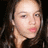 Pixelated close-up of a person with light skin, dark eyes, and long dark hair, wearing a dark top against a dark background.