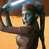 Static pixelated image of a humanoid figure with metallic blue head and dark body holding a large cylindrical object against a gradient background.