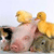 A pixelated image shows a light pink pig lying on its back with two yellow ducklings perched on its belly against a pale blue background.