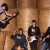 A pixelated group of four figures in dark clothing against a brown wooden background, with one figure holding a guitar-like object.