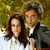 Two people with brown hair stand close together against a blurred green and yellow background, one wearing dark clothing over white.
