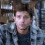 A pixelated image of a person with short dark hair wearing a blue jacket over a dark shirt, standing against a blurred background.