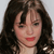 A pixelated close-up of a person with long dark brown hair, bangs, and red lips against a light gray background.