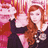A woman with long red hair in a black dress stands against a pink and orange striped background, her right hand raised near her head.