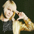 Pixelated image of a person with light hair holding a microphone against a dark background.