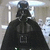 Static pixelated image of a figure in black armor and cape with helmet, standing against light gray background.