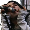 A person in a light gray jacket holds a black camera with a large lens up to their face, positioned in front of a blurred background.