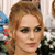 A pixelated close-up of a person with light brown hair styled in an updo, wearing dark eyeliner and red lipstick, with a blurred background containing blue, white, and green elements.