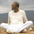 A pixelated figure in white clothing sits cross-legged on a sandy surface with a blurred background of water and sky.