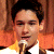A pixelated close-up of a person with dark hair holding a microphone, wearing an orange garment against a brown background.