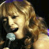 A woman with light brown hair sings into a microphone, wearing a dark sleeveless top under stage lighting.