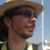 Pixelated profile view of a person wearing a wide-brimmed straw hat, dark sunglasses, and a light-colored shirt against a bright sky background.
