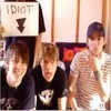 Three people pose together with one holding a sign reading 'IDIOT' and a downward arrow.