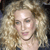 Pixelated close-up of a person with light brown curly hair, fair skin, and a neutral expression, wearing a dark top against a blurred background.