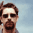 Pixelated image of a person with dark hair and a beard, wearing dark sunglasses, holding a phone to their ear against a light blue background.