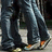 Pixelated static image of two figures in dark clothing with light-colored accents, standing on a textured surface under dim lighting.