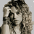 A grayscale image of a woman with curly hair holding her hand near her head, wearing a sleeveless top and standing before a microphone.