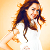 A woman with long brown hair wearing a white dress stands against a light orange background with one hand on her hip.